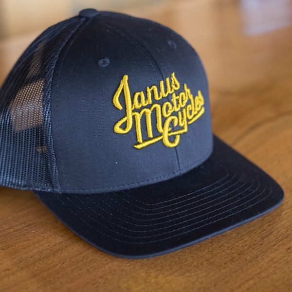 Navy blue Janus Motorcycles cap with yellow embroidery on a wooden surface.