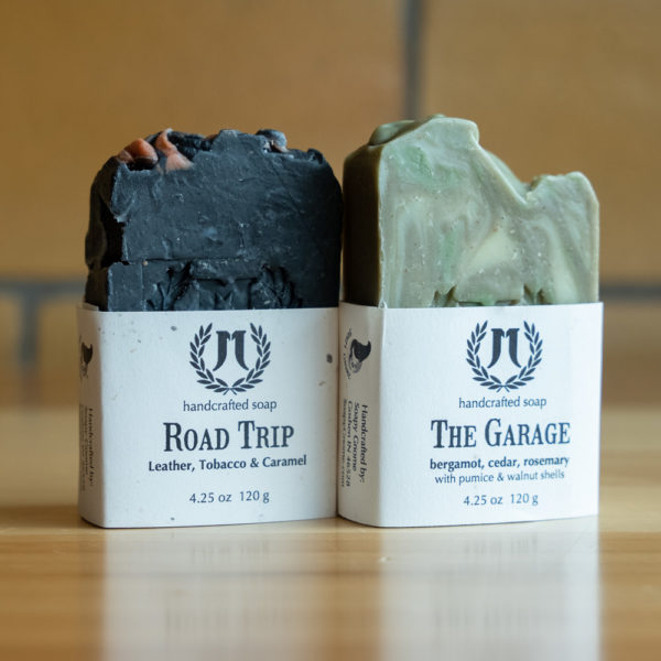 Two handcrafted soaps labeled Road Trip and The Garage on wooden surface.