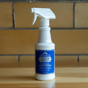 Spray bottle of Obenauf's Water Shield waterproofing on wooden surface against brick background.