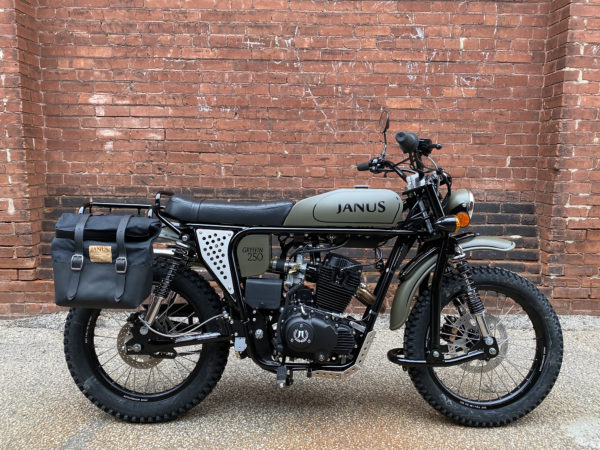 Vintage-style Janus motorcycle with side bag parked in front of a brick wall.