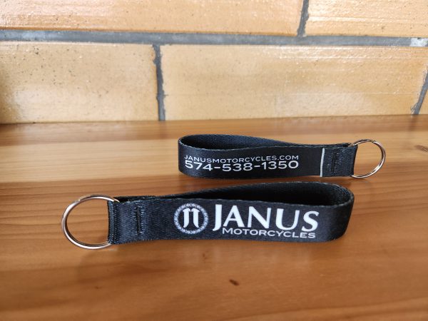 Black keychain with Janus Motorcycles logo on wooden table.