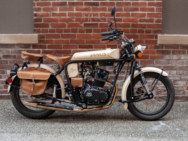 Cream-colored vintage Janus motorcycle with leather saddlebags parked by a brick wall.