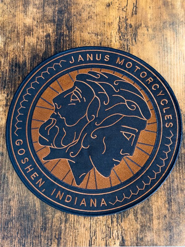 Round Janus Motorcycles branded coaster with a two-faced profile on a wooden table.