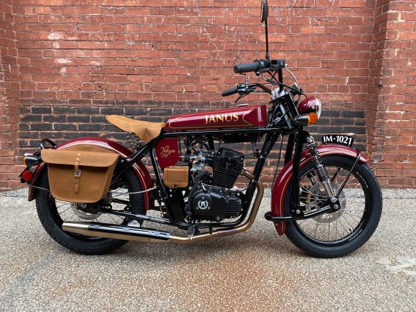 Classic red and maroon Janus motorcycle with tan saddlebags parked against a brick wall.
